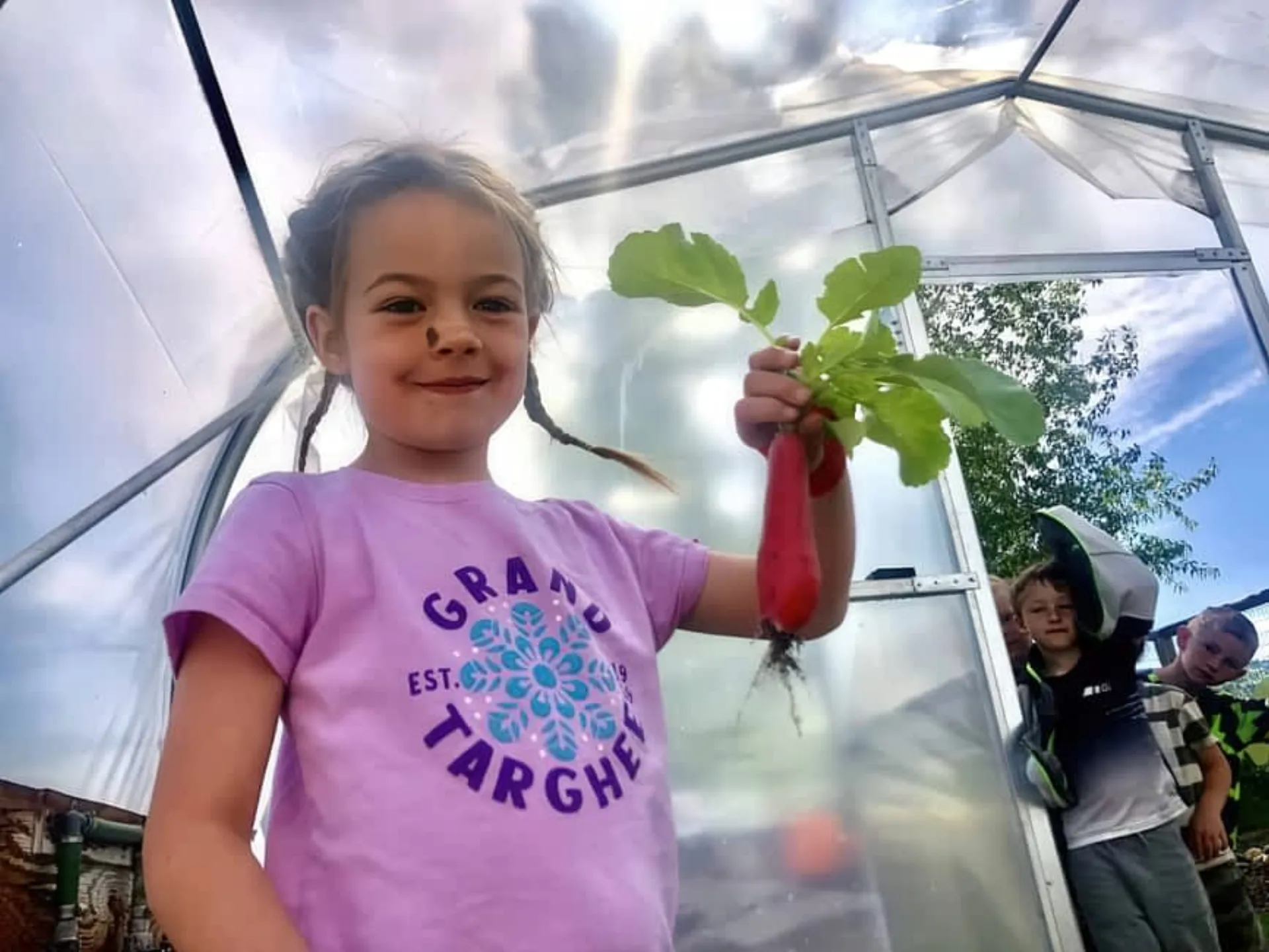 Young girl proudly holding a freshly picked radish in a community greenhouse supported by the Community Foundation